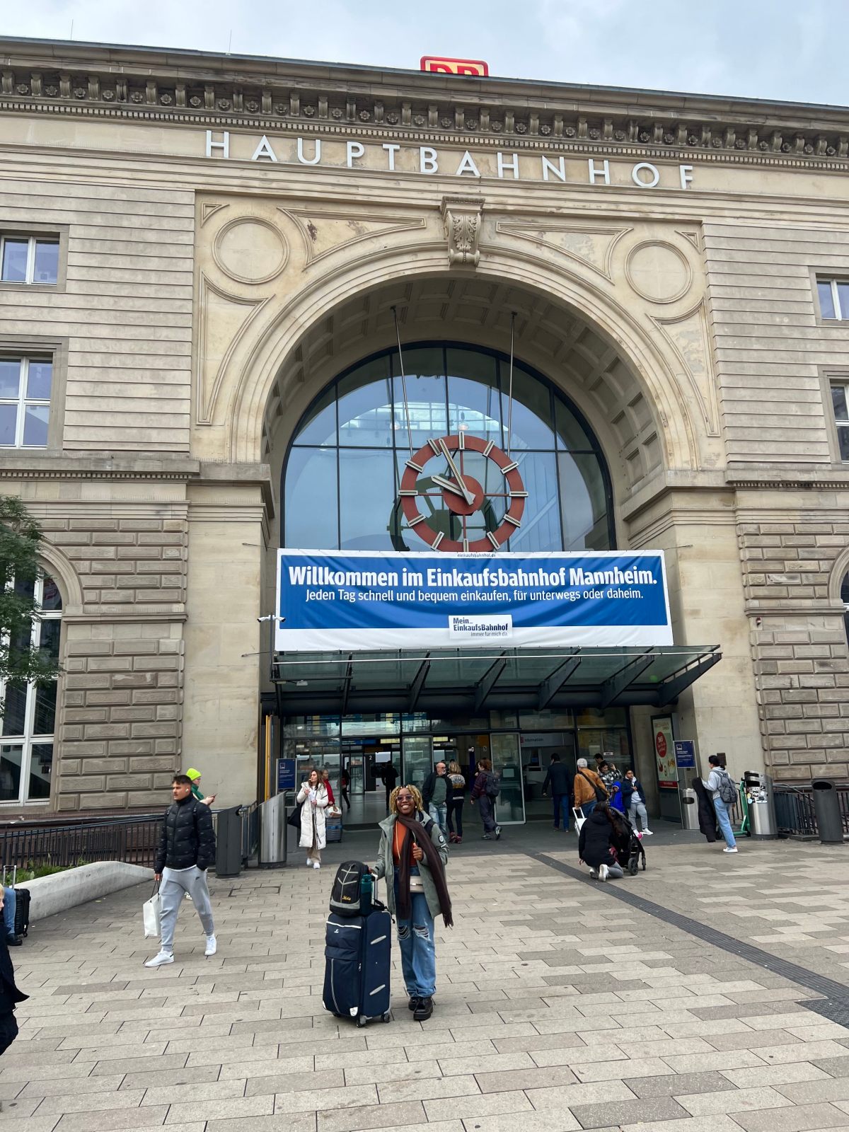 A smiling person standing with their luggage in front of Mannheim Hauptbahnhof, poised to embark on a journey to Berlin.