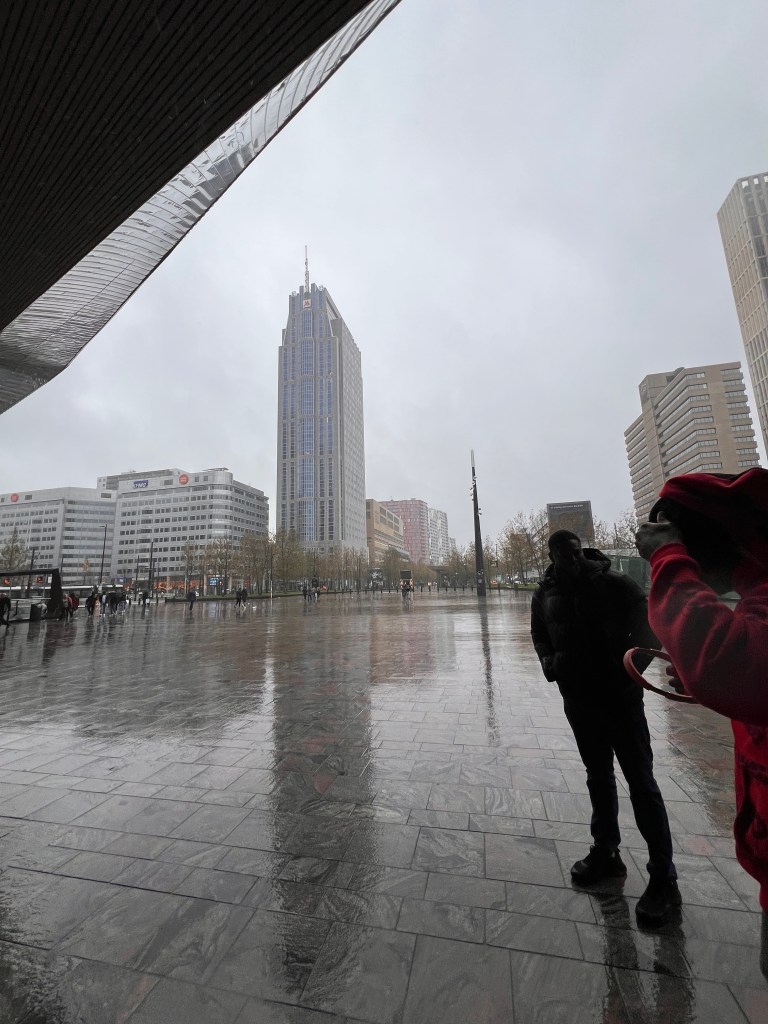 View from under a shelter looking out at Rotterdam Centraal Station on a rainy day, with Timmy standing in the foreground and city buildings in the distance