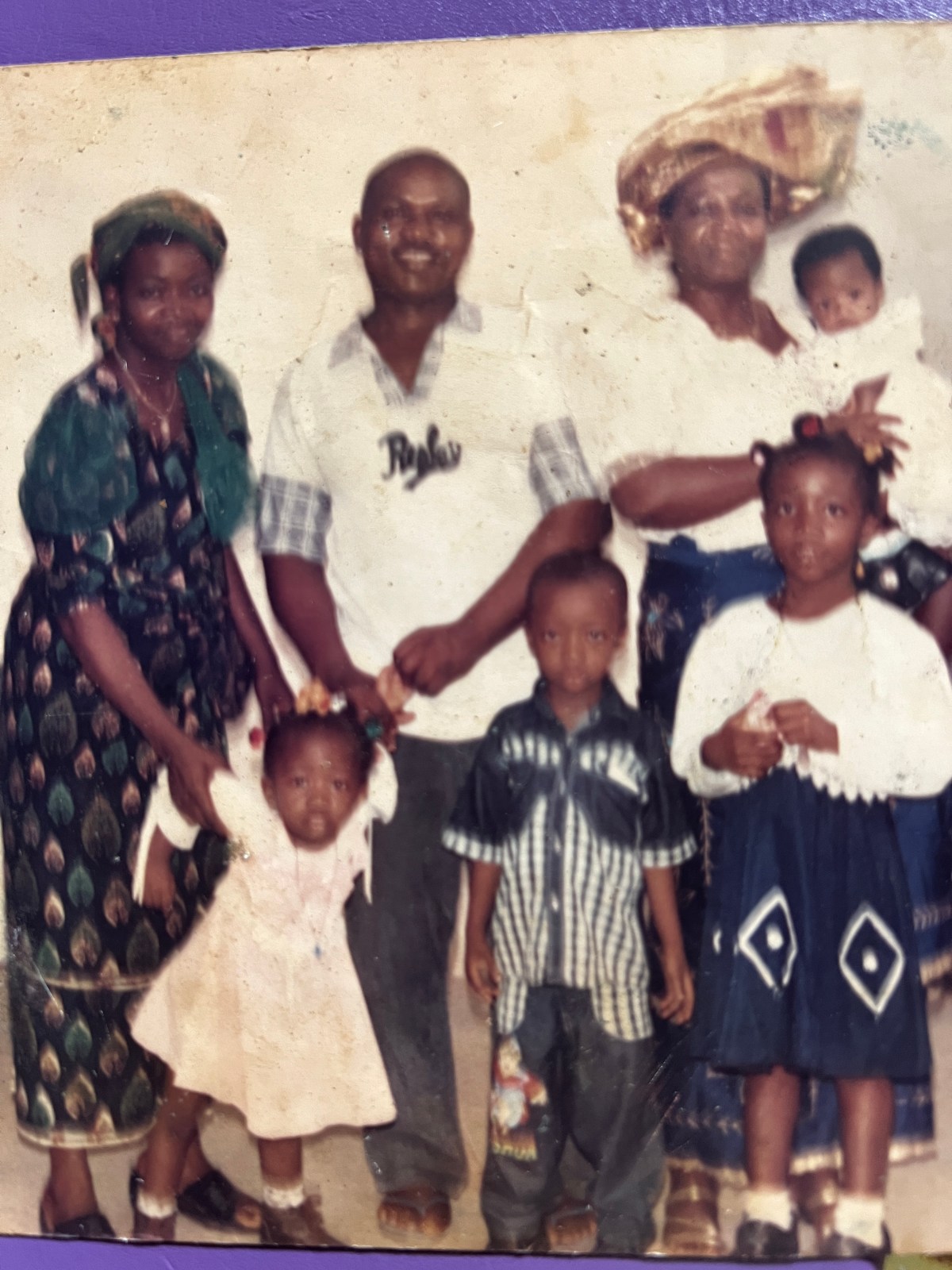 Family photo at a church after a baptism. From left to right: A woman in a green patterned outfit holding a small girl, a man in a white shirt, a woman in white holding a baby, and two young children standing in front.