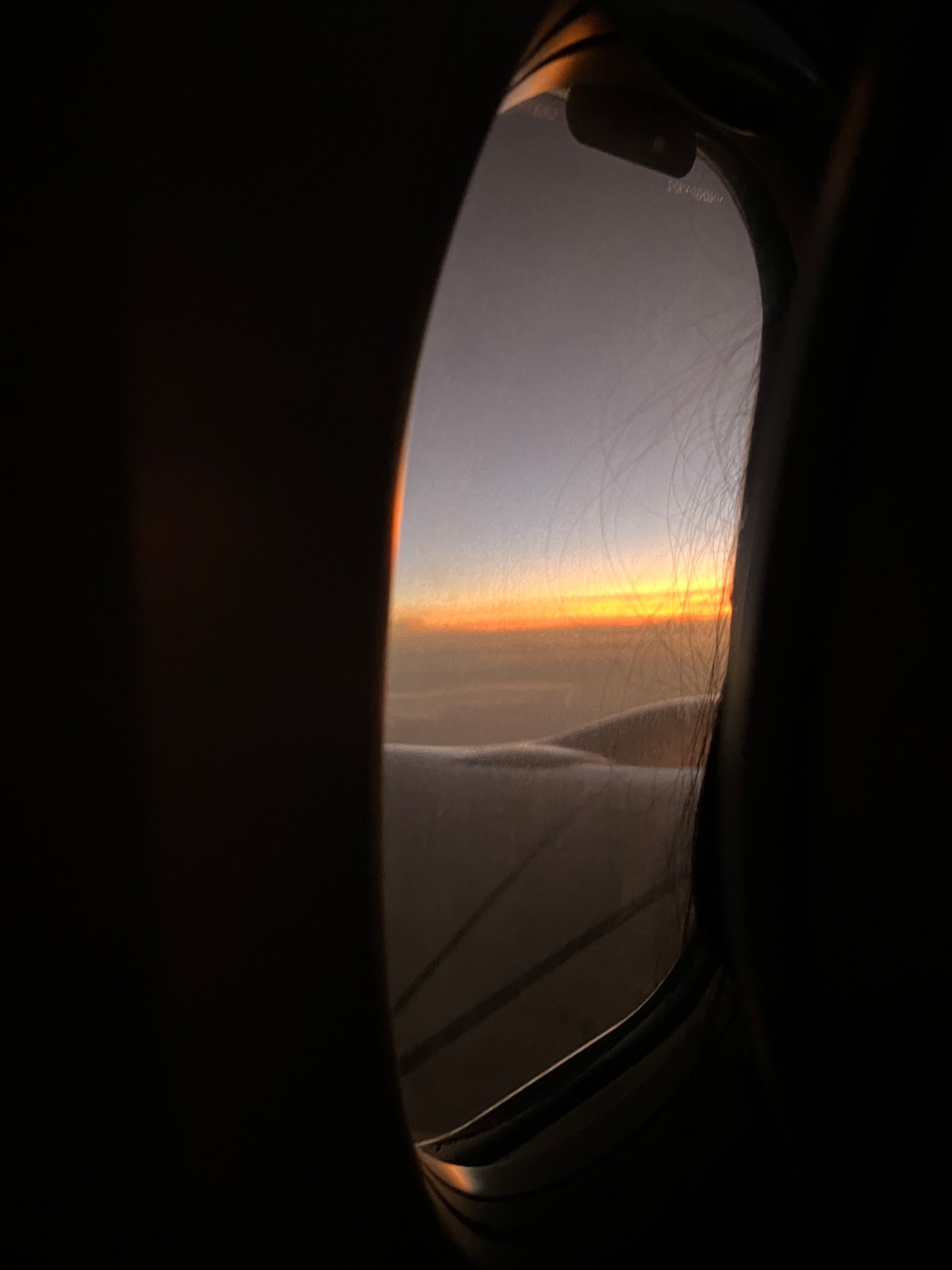 A view through Kenya Airways aircraft's porthole. The scene outside captures a serene horizon with soft hues of orange, yellow, and blue blending at sunrise